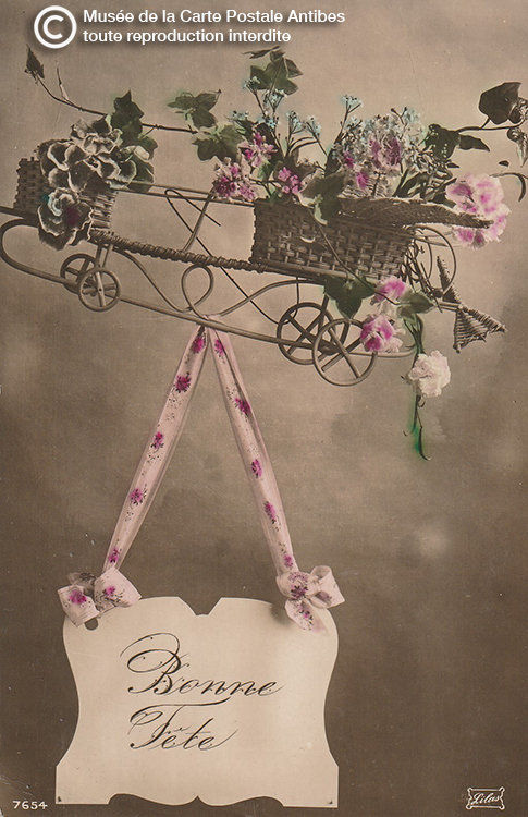 Carte postale aéroplane en osier tressé transportant le panneau « Bonne Fête ». Type de cartes pouvant être utilisée en toutes circonstances. On distingue les ailes, les roues, l’hélice de la maquette censée représenter un avion. Editeur Lilas phototypie colorisée main, N°7654 (avant 1914).